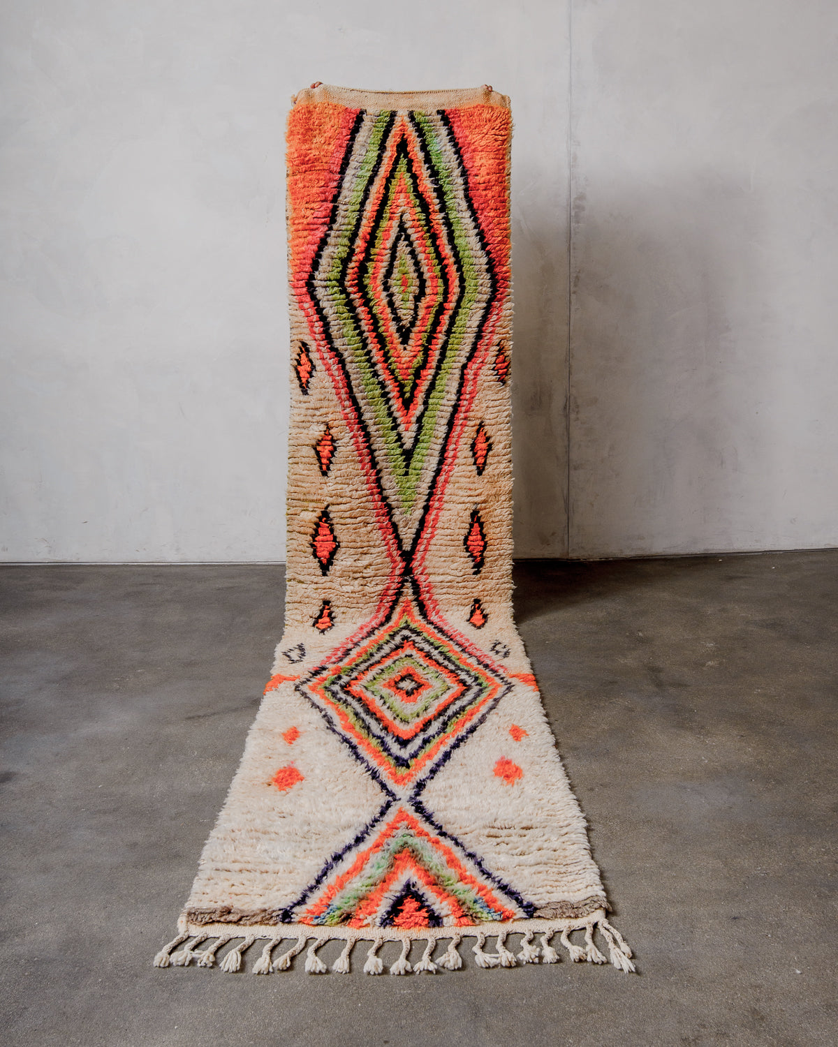 Berberlin vintage runner rug made of cream and brightly coloured wool, being held up in front of a white wall and concrete floor.